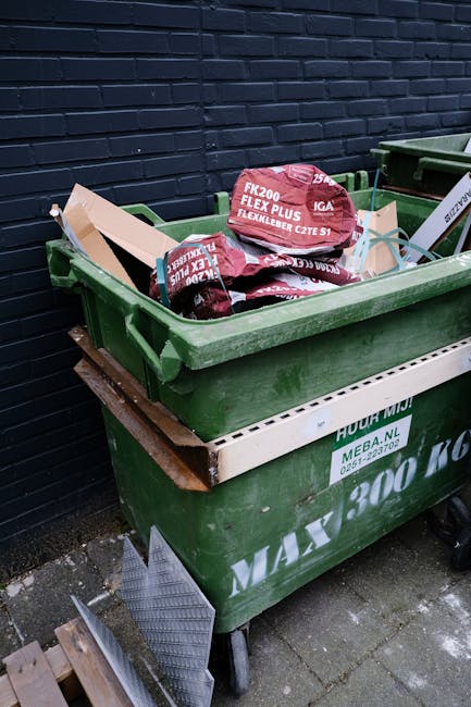 A black wheeled rubbish bin marked with the name 'St. John's' is positioned on a paved sidewalk beside a residential street at night. The bin is partially open and filled with various discarded materi