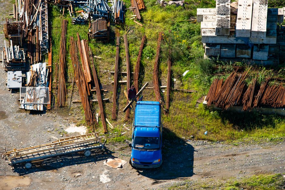 Aerial view of a large industrial scrapyard or waste disposal site on a clear day, featuring multiple piles of scrap metal and discarded vehicle parts. In the foreground, a row of compacted metal car 